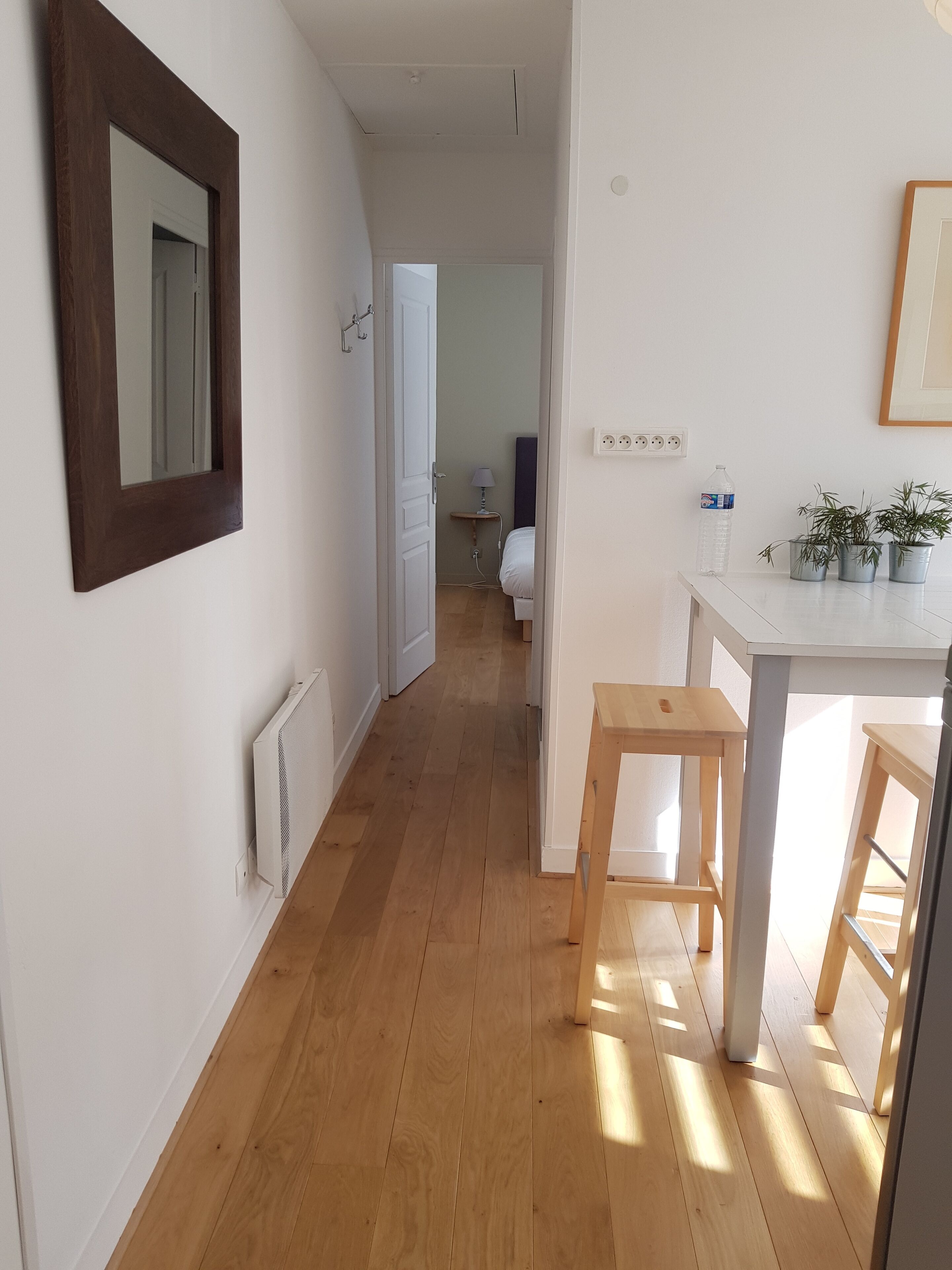 A narrow hallway with wooden flooring, a white radiator, a mirror, and a door leading to a bedroom with a bed and a nightstand.
