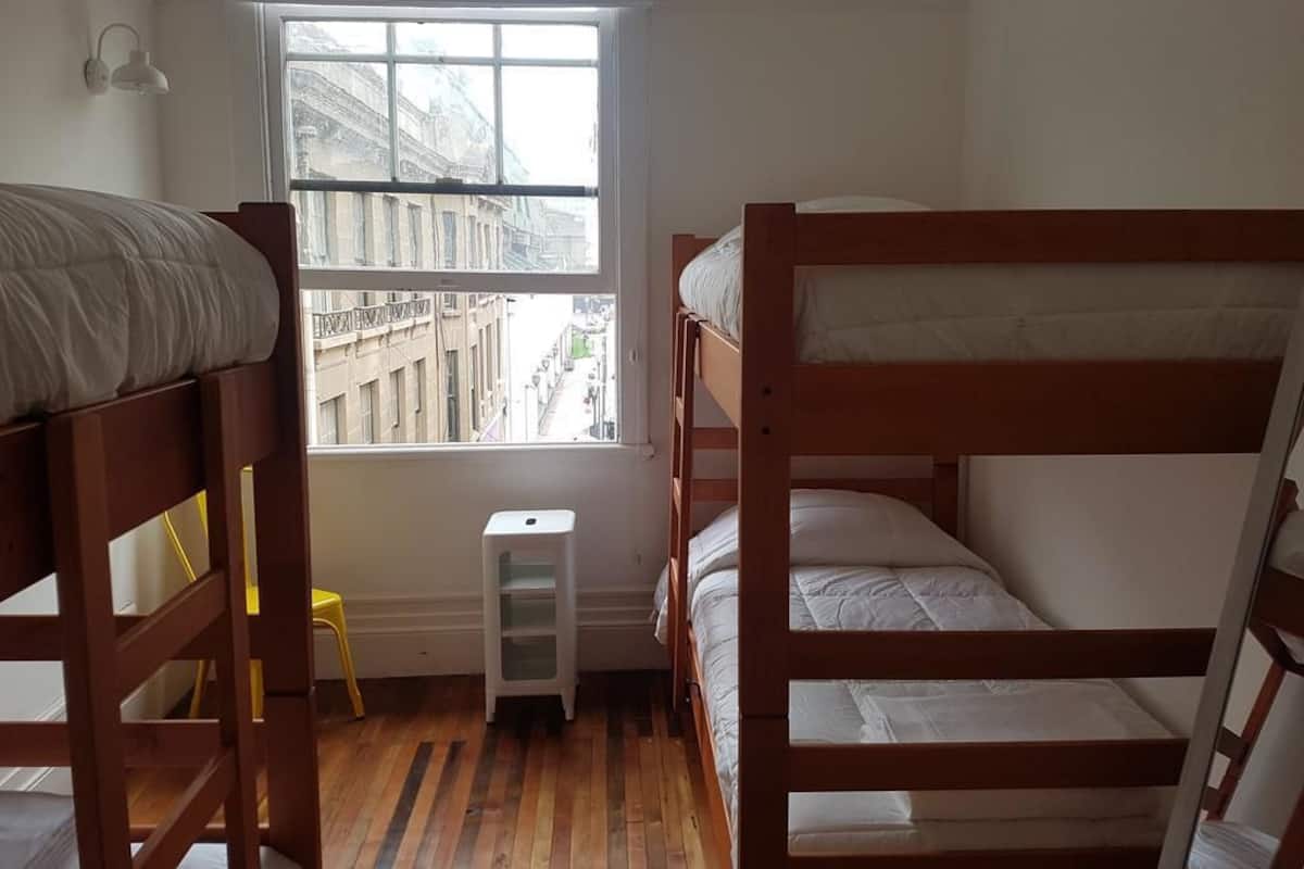 Quarto com beliches, janela com vista para edifícios, um banquinho pequeno e um armário branco.