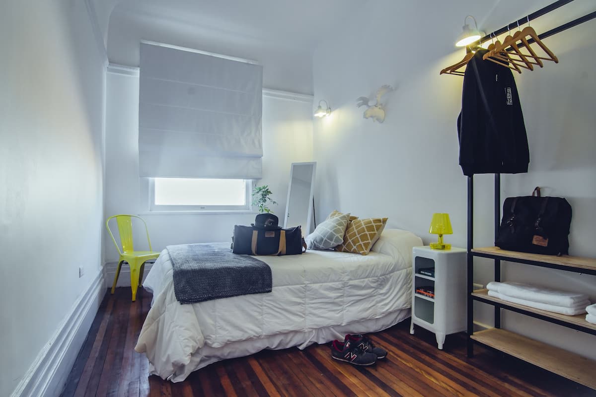 Quarto compacto com cama, cadeira, mesa de cabeceira, luminária, espelho, guarda-roupa e prateleira com toalhas.
