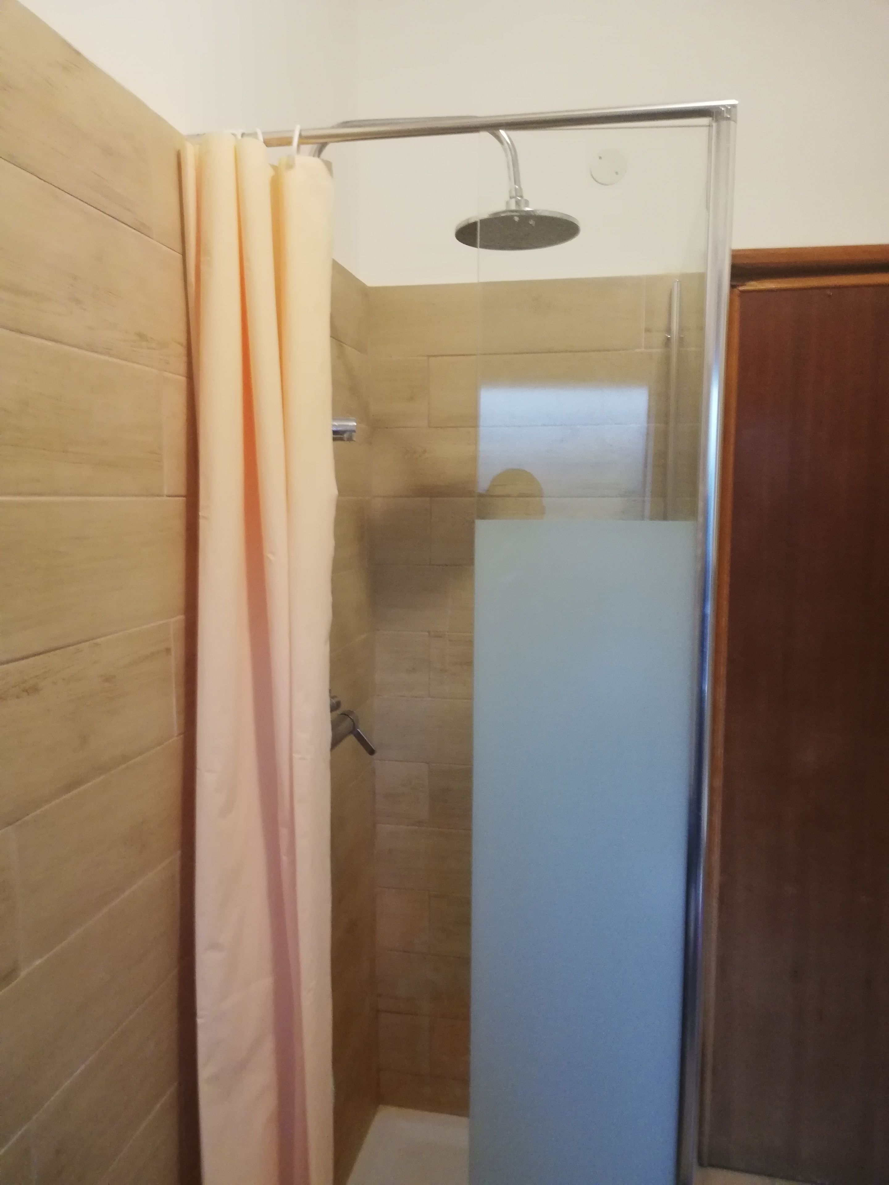 Ducha con mampara de vidrio, paredes de azulejos color beige y marco de puerta de madera.