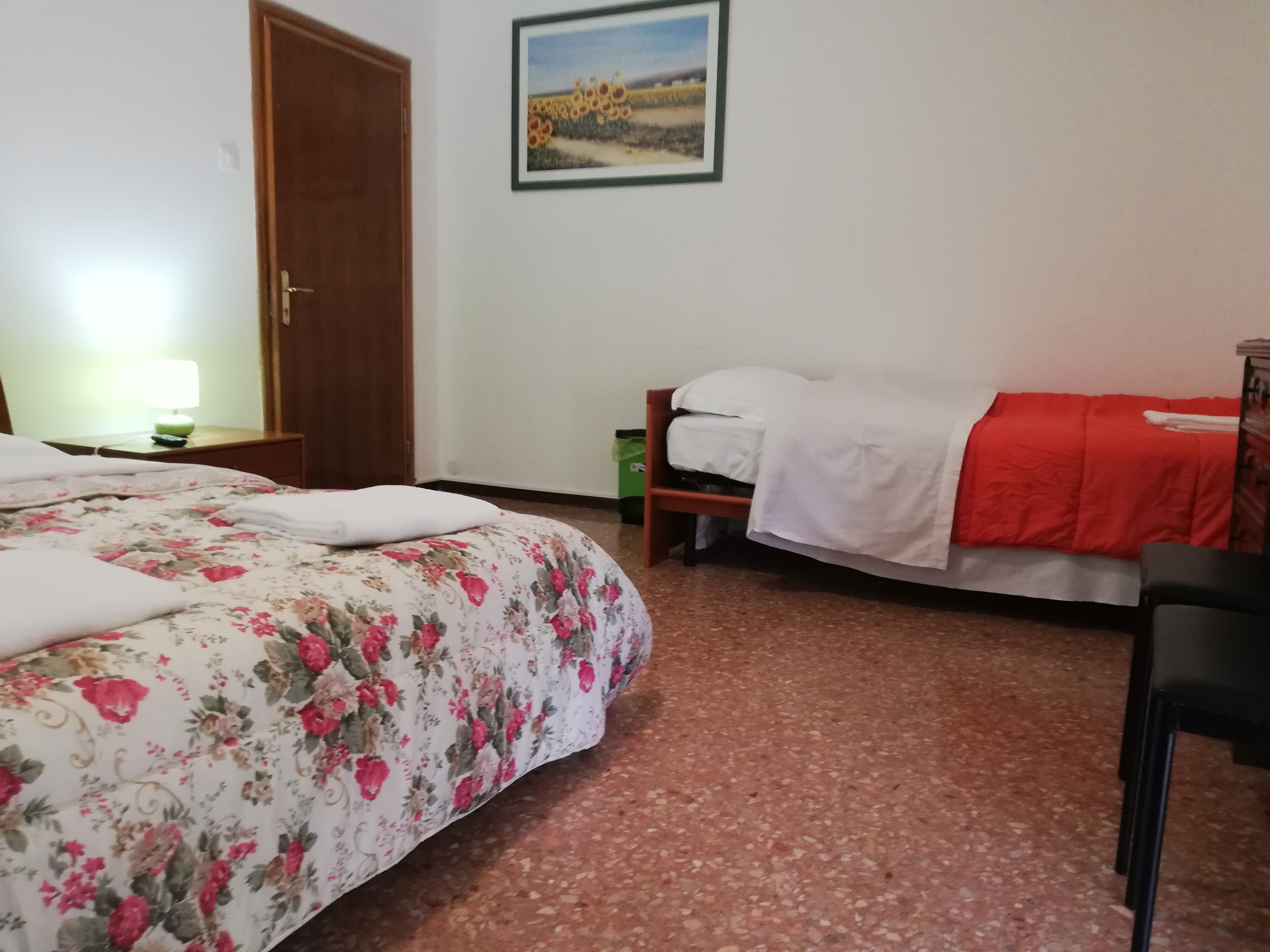 Una habitación con edredón estampado, una cama blanca, una manta roja, un cubo de basura verde y un cuadro enmarcado en la pared.