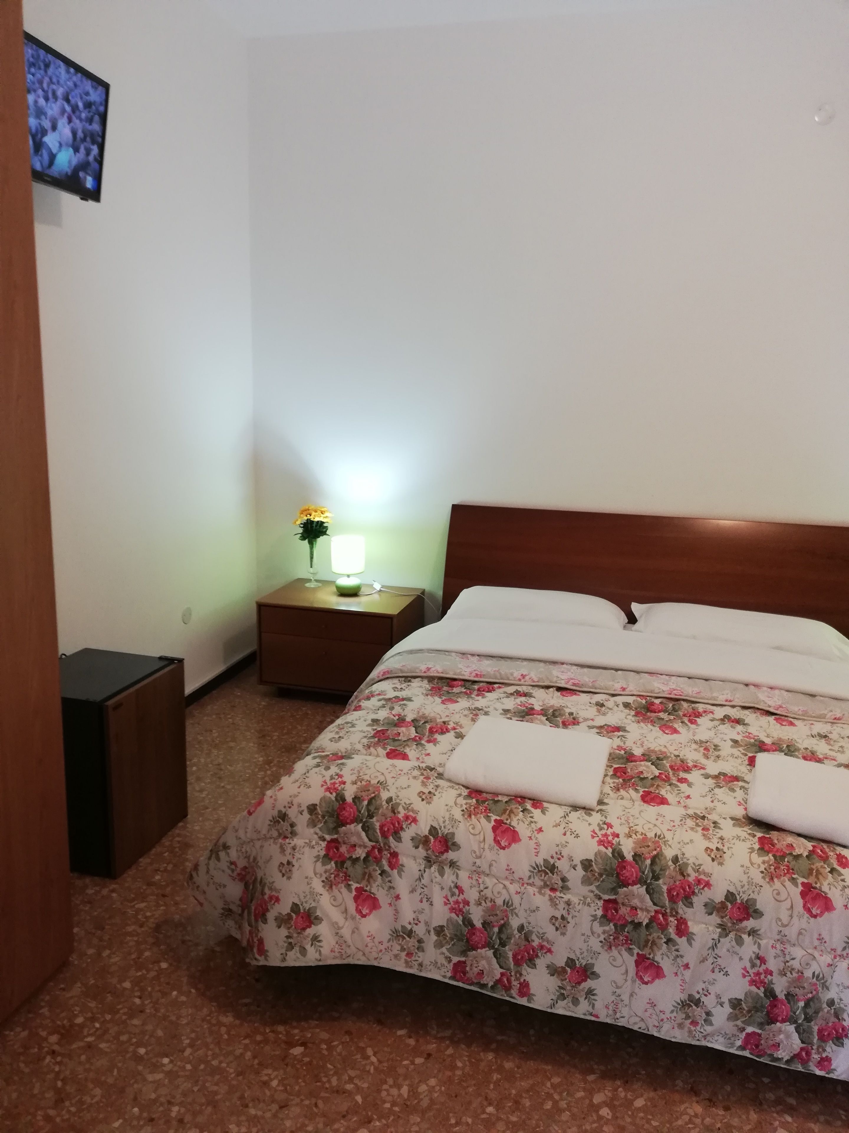 Un dormitorio con colcha estampada, cabecero de madera, mesita de noche con lámpara y televisor montado en la pared.