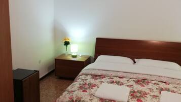 A bedroom with a floral bedspread, a wooden headboard, a bedside table with a lamp, and a TV mounted on the wall.