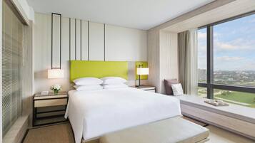 A modern hotel room with a large bed, a bedside table with a lamp, a bench, and a view of the city through large windows.