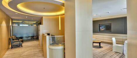 Un vestíbulo de hotel moderno con suelo de madera, una recepción, una zona de estar y un televisor montado en la pared.
