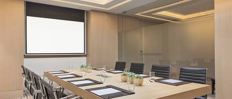 A conference room with a long table, chairs, a projector screen, and potted plants.