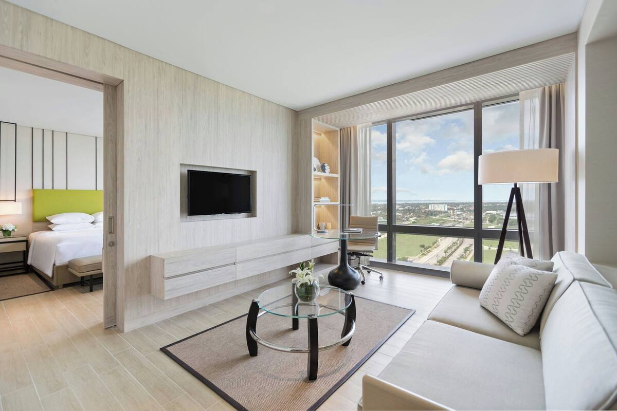 A modern hotel room with a flat-screen TV mounted on the wall, a sofa with a view of the city, a glass coffee table, and a large window with a view of the cityscape.