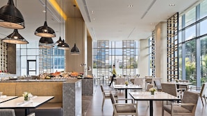 Um restaurante moderno com pé-direito alto, iluminação pendente e grandes janelas com vista para a cidade.