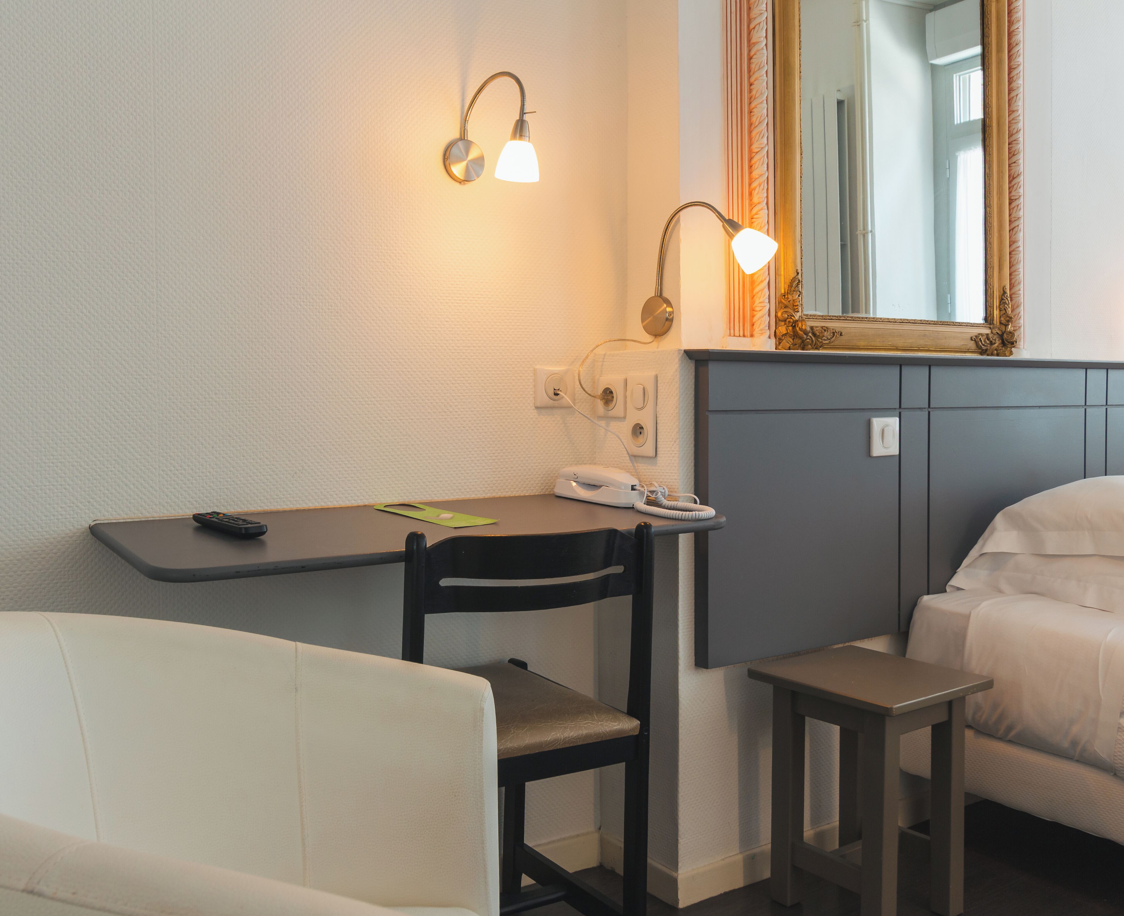 A hotel room with a bed, a desk with a telephone, a chair, a small table, and a wall-mounted lamp.