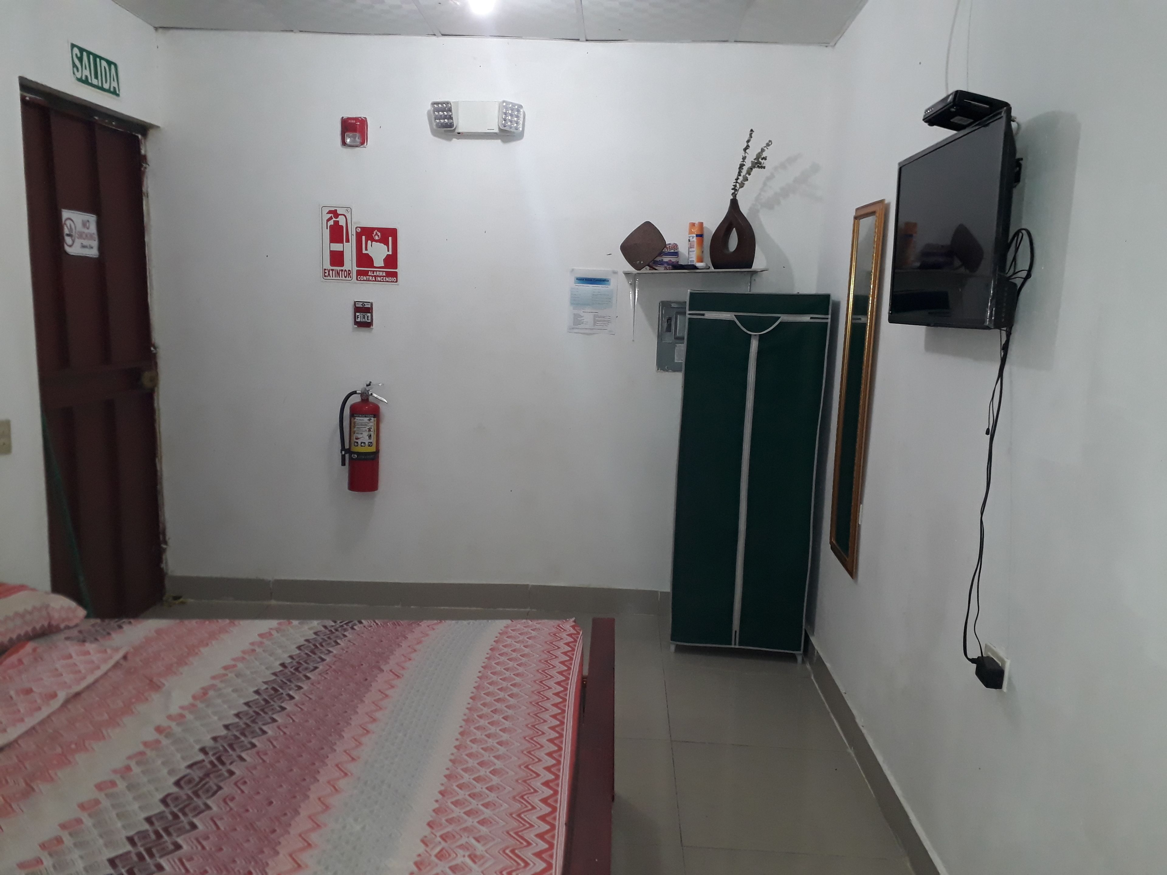 Una habitación con una cama, extintor, detector de incendios, televisor y armario.