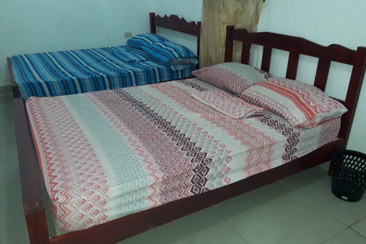 A bed with a patterned bedspread and pillows, a wooden headboard, and a black trash bin beside it.