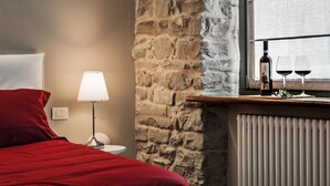 A bedroom with a stone wall, a bed with red bedding, a bedside table with a lamp, and a window with two wine glasses and a bottle. - Albergo del Bramante (Roccaverano)