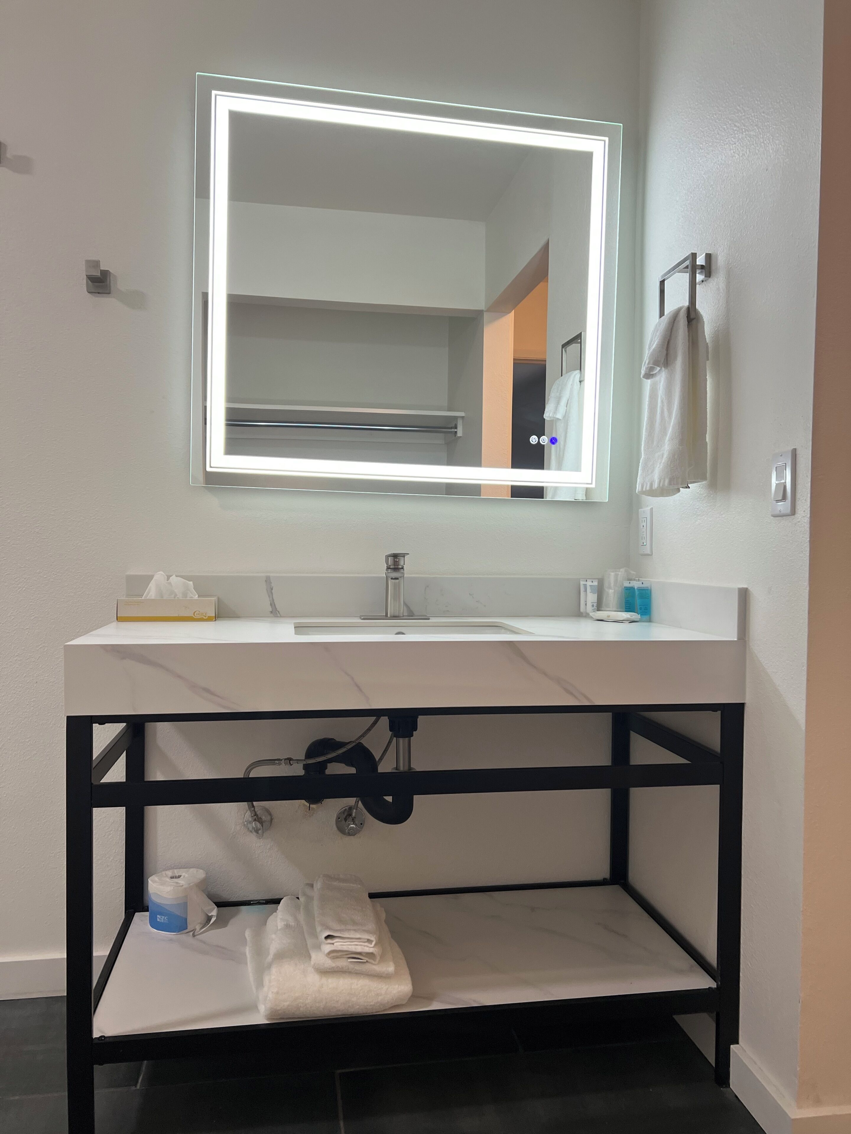 Un baño con un tocador blanco, un espejo con luz, un perchero y una toalla en el lavabo.