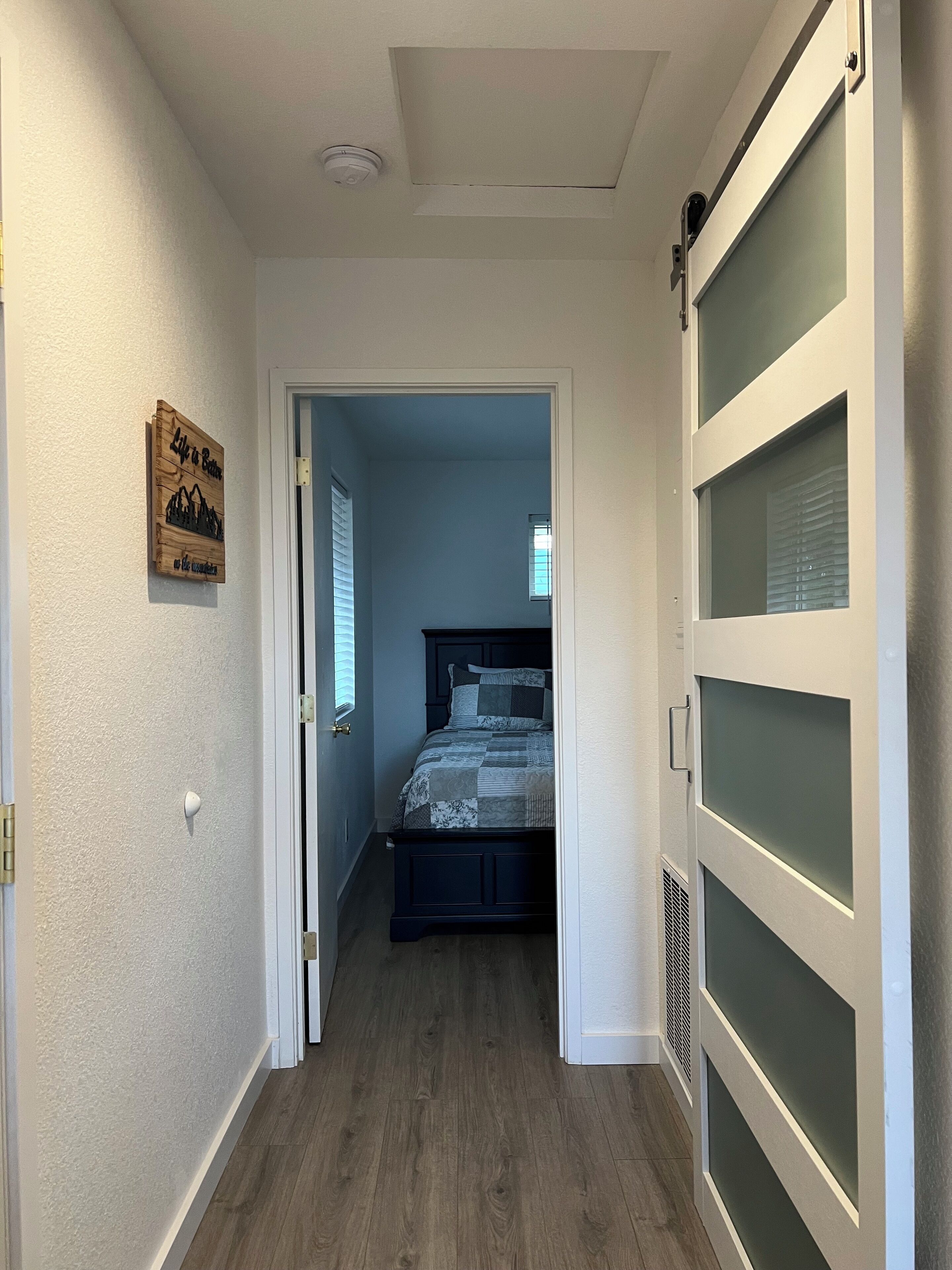 A narrow hallway leading to a bedroom with a bed, a wooden sign on the wall, and a window with blinds.