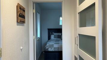 A narrow hallway leading to a bedroom with a bed, a wooden sign on the wall, and a window with blinds.
