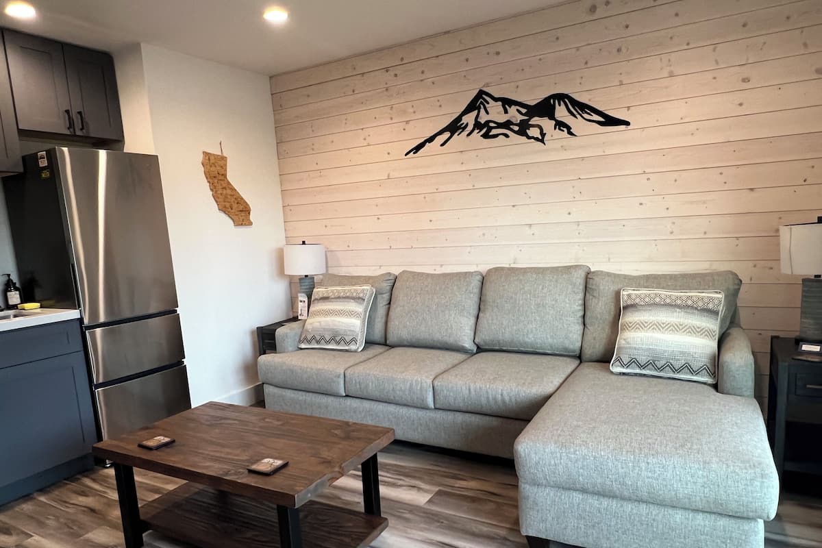 Una sala moderna con un sofá modular, una mesa de centro de madera, una cocina con refrigerador de acero inoxidable y una pared de madera con diseño de montaña.