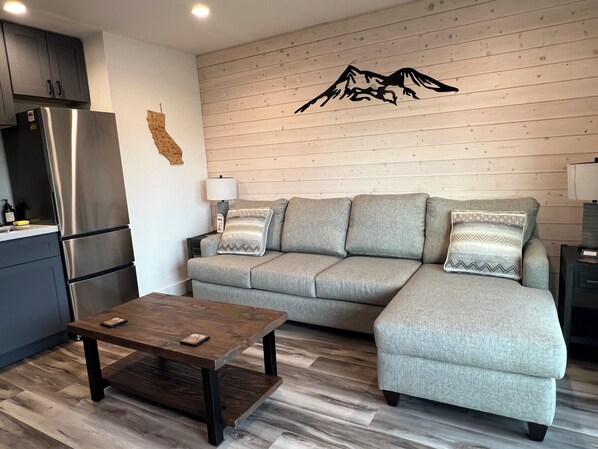A modern living room with a sectional sofa, a wooden coffee table, a kitchen area with a stainless steel refrigerator, and a wooden wall with a mountain design. - Lake Shastina Golf Resort (Weed)