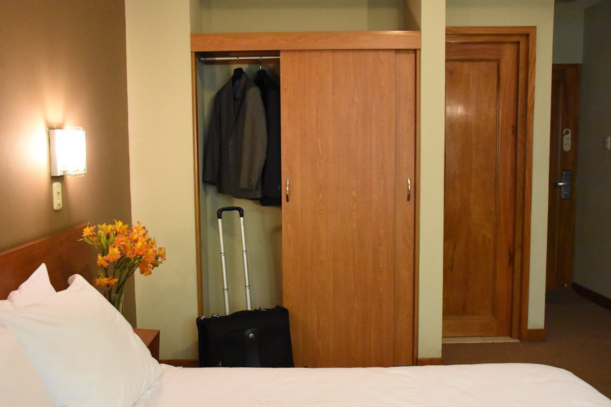 Una habitación de hotel con una cama, una mesita de noche con un florero y un armario de madera, además de una maleta.