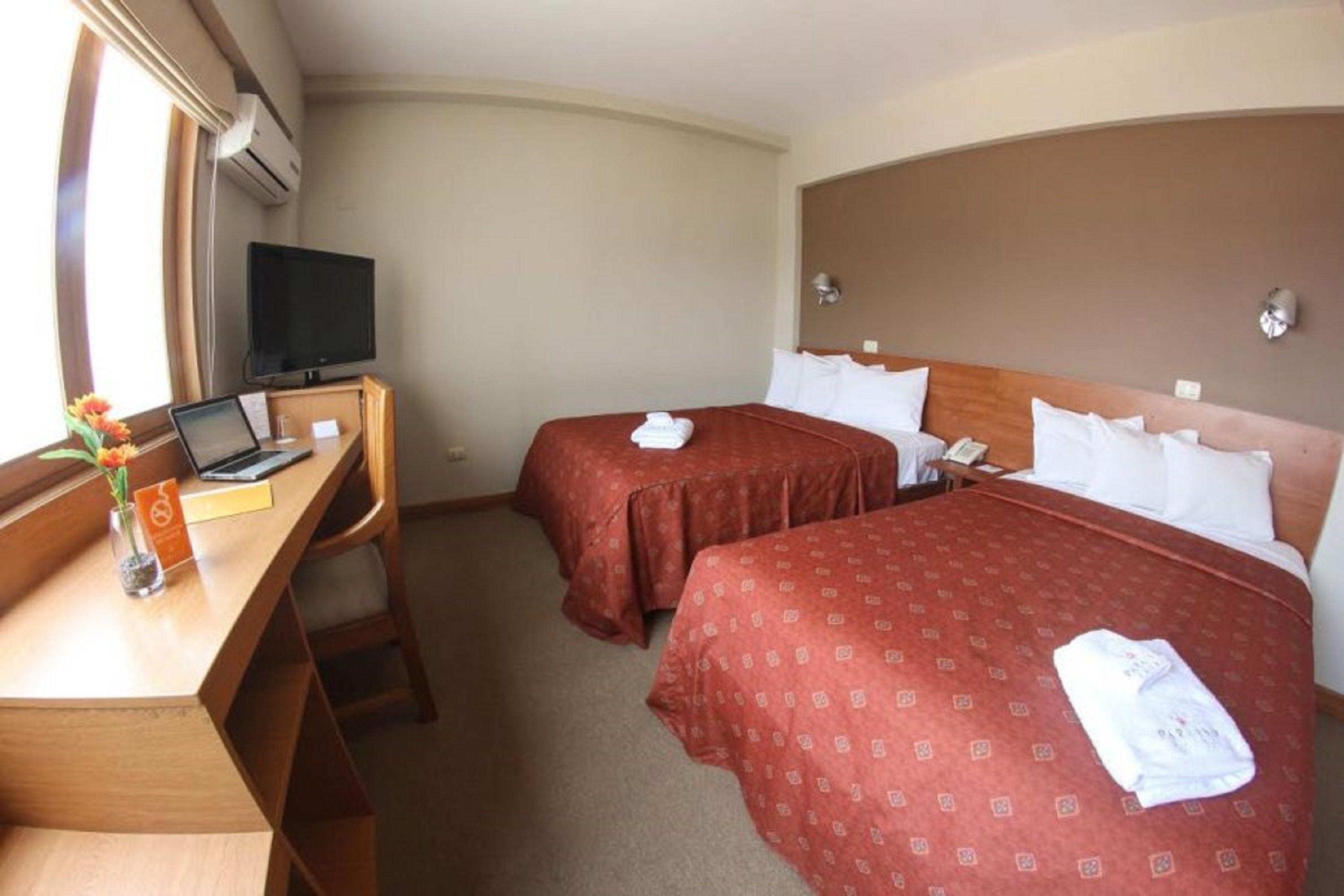 A hotel room with two beds, a desk with a laptop, a television, and a window with curtains.