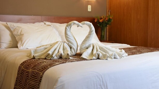 A hotel room with a bed featuring a towel arrangement in the shape of swans, a patterned throw blanket, and a vase with flowers on the nightstand. - Hotel Paraiso Trujillo (Trujillo)
