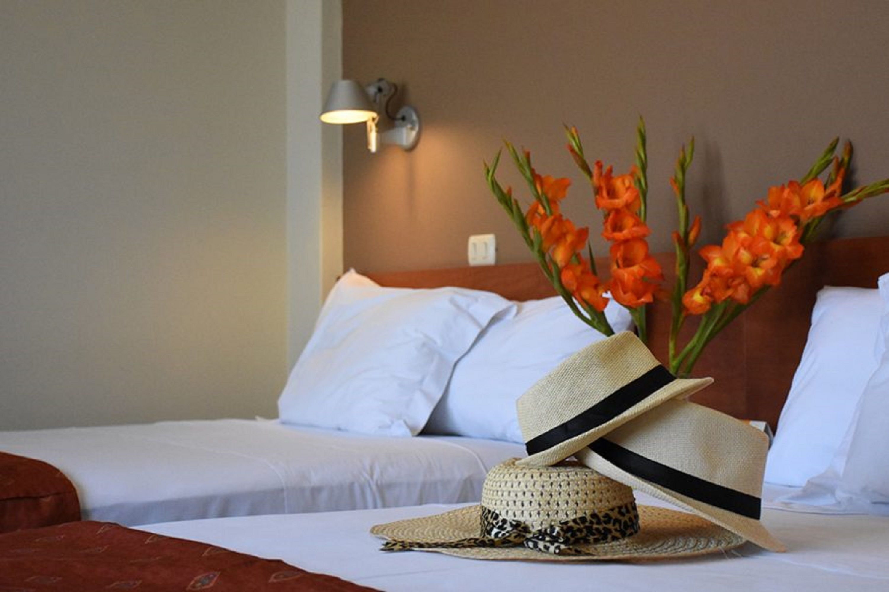 Habitación de hotel con dos camas, una lámpara de pared y un florero con flores anaranjadas.