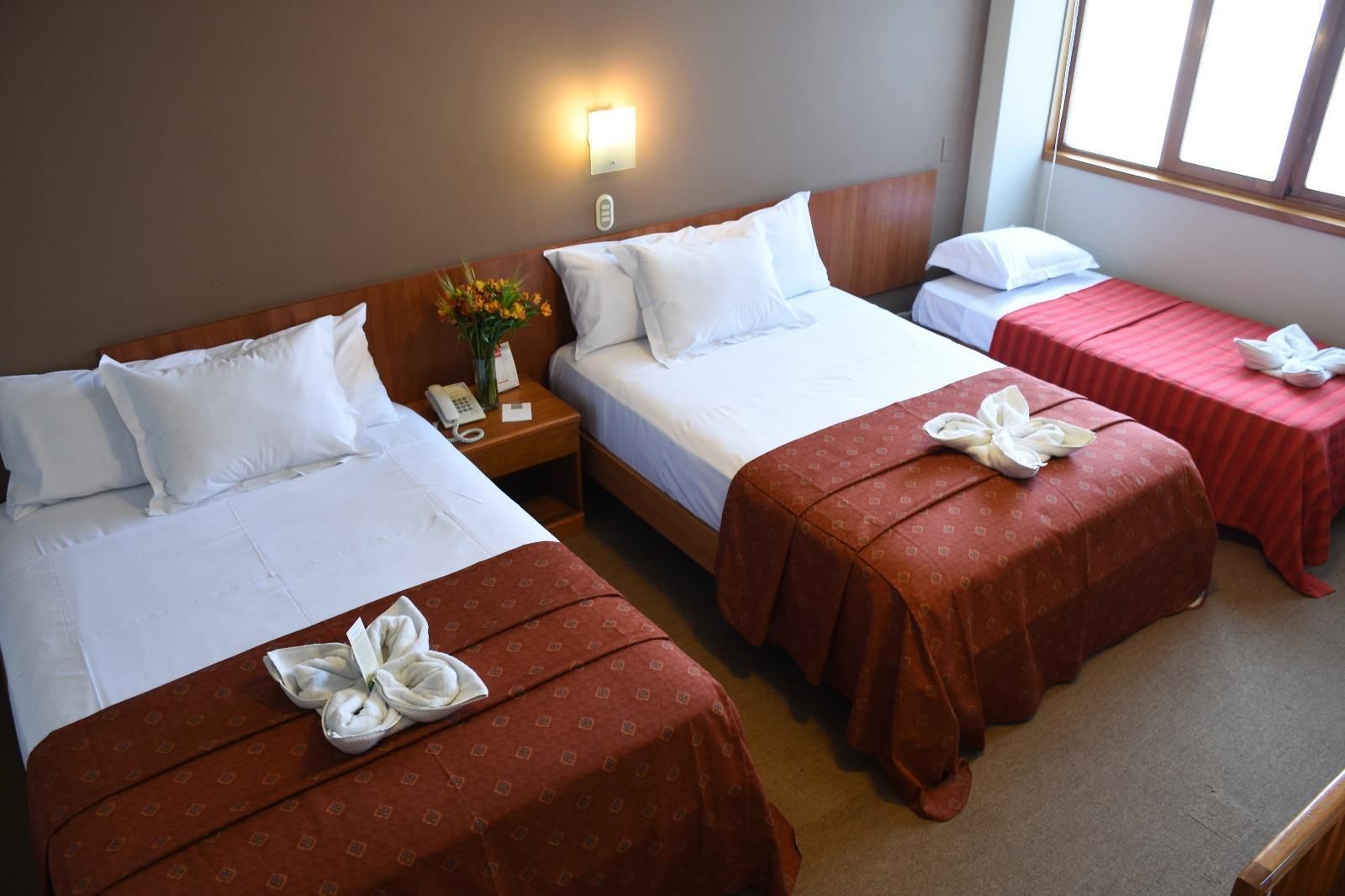 Habitación de hotel con tres camas, cada una con sábanas blancas y colchas marrones. A la izquierda, hay una mesita con un jarrón de flores y un teléfono. A la derecha, la habitación tiene una ventana.