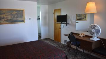 A hotel room with a bed, desk, chair, television, fan, and artwork on the walls.