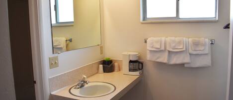 Un baño con lavamanos, espejo y ventana. Hay toallas colgadas en la pared y una cafetera sobre la encimera.