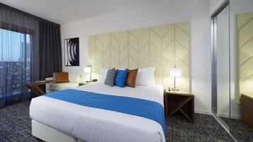 A hotel room with a large bed, two bedside tables, a nightstand, a chair, a window with sheer curtains, and a wall-mounted artwork.