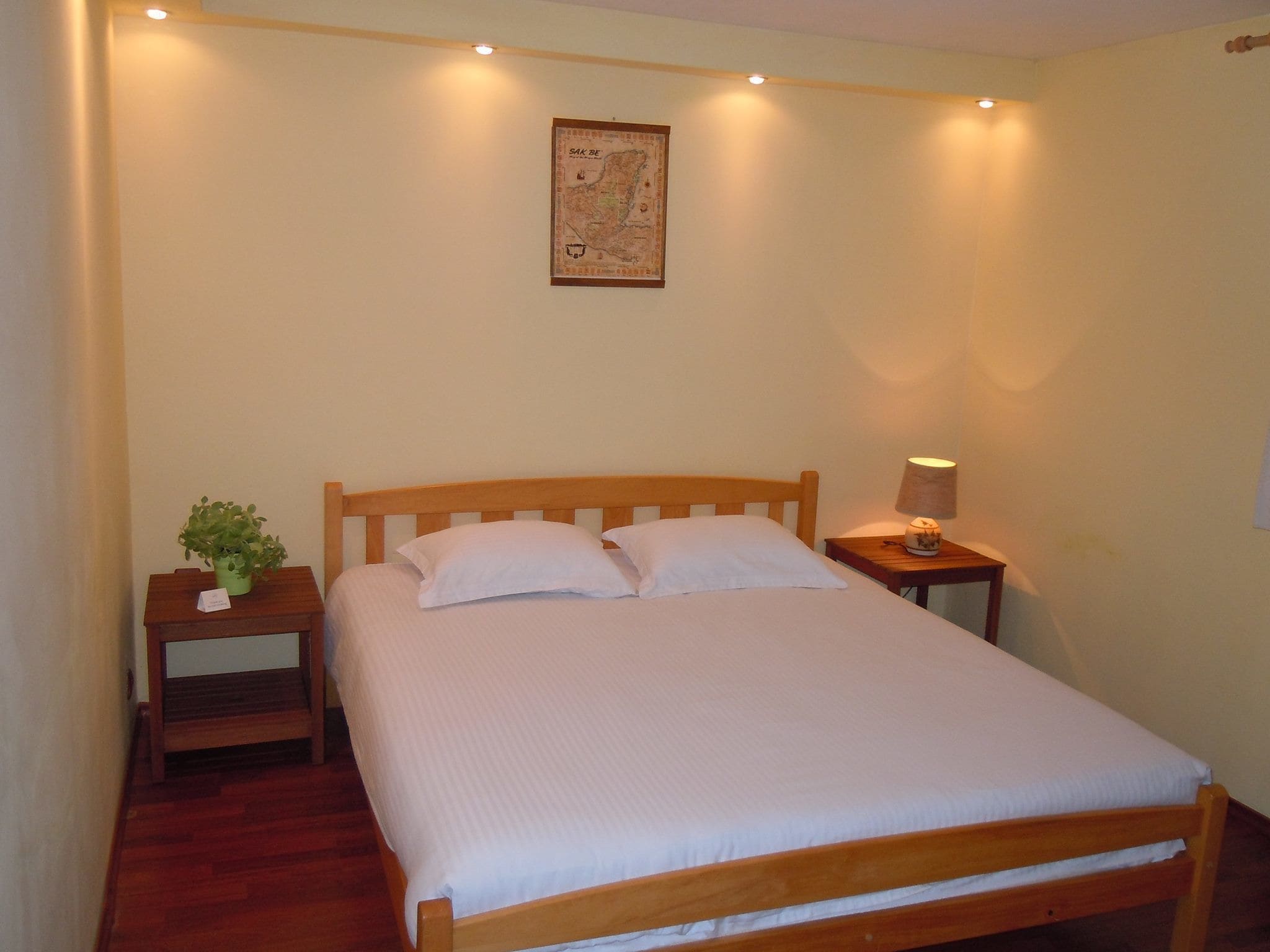Una cama bien hecha con ropa de cama blanca en una habitación con muebles de madera, una mesita de noche con una lámpara y un mapa en la pared.
