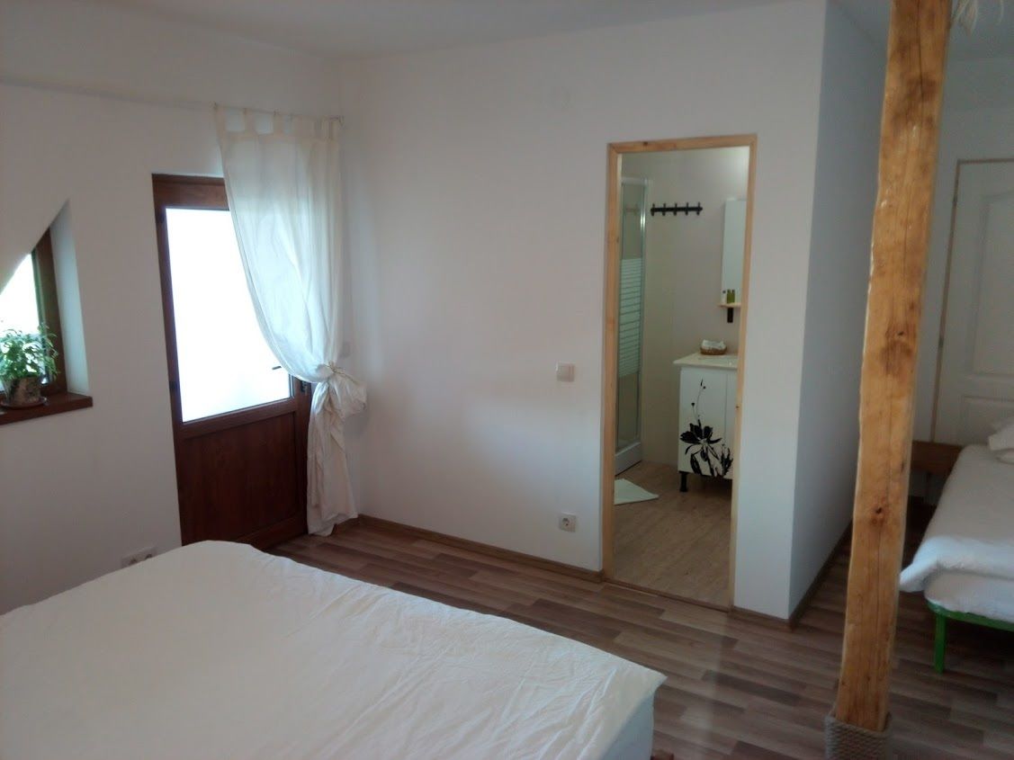 Quarto com cama, janela com cortinas, piso de madeira e espelho refletindo um banheiro.