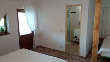 Quarto com cama, janela com cortinas, piso de madeira e espelho refletindo um banheiro.