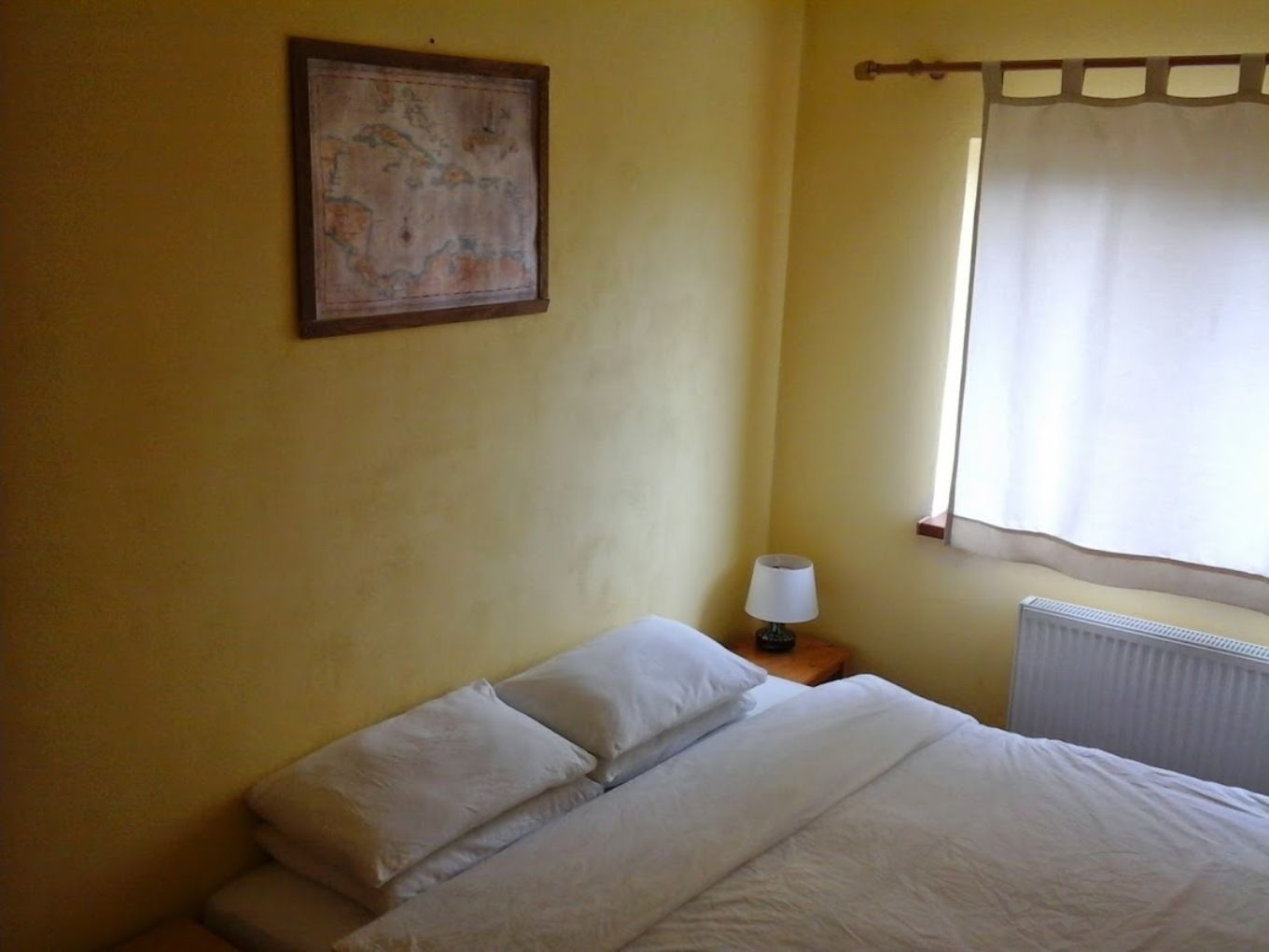 A bedroom with a bed, a nightstand with a lamp, a radiator, and a map on the wall.
