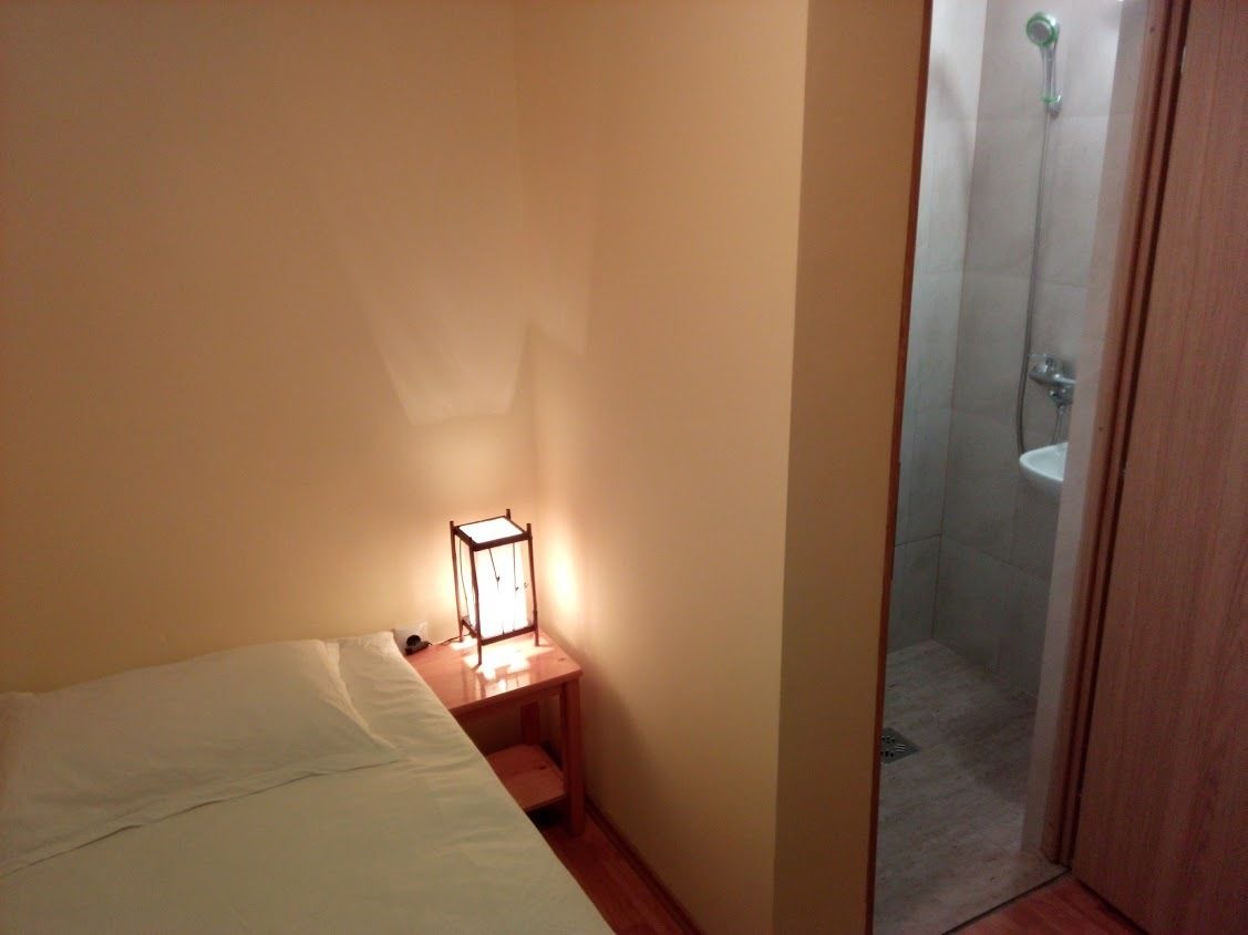 Una habitación pequeña y con poca luz, con una cama individual, una mesita de noche con una lámpara, y un rincón de ducha visible a través de una puerta de cristal.