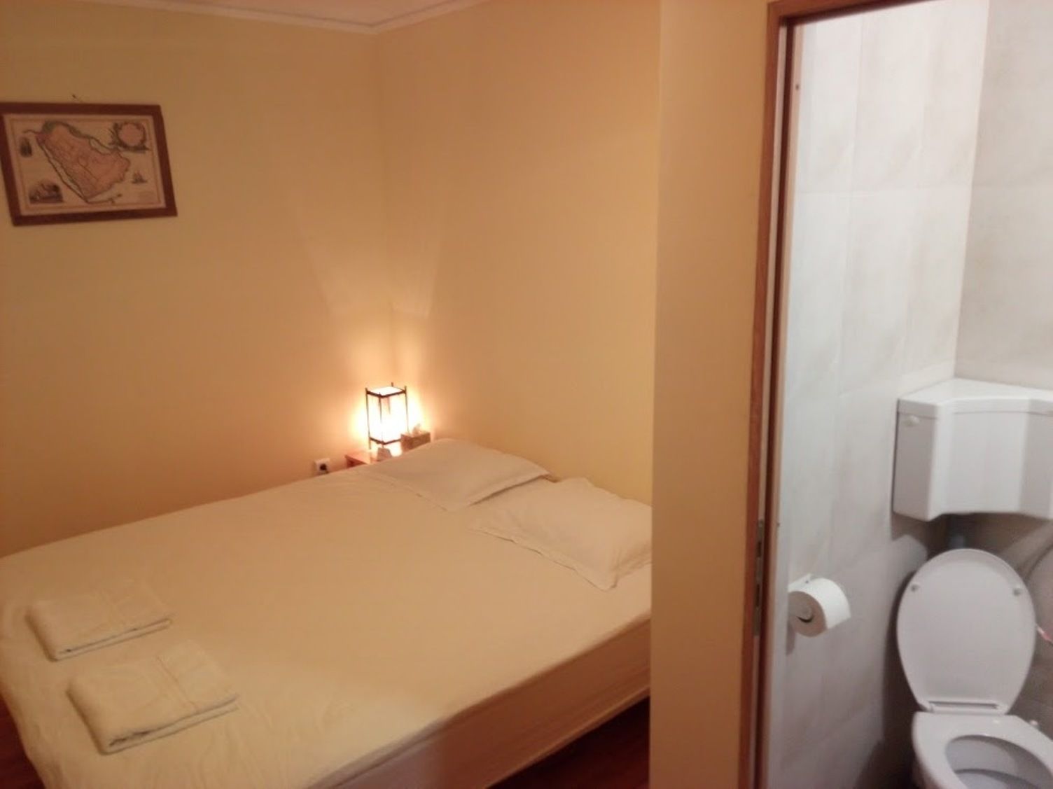 Habitación pequeña con cama individual, mesita de noche, lámpara y un inodoro visible a través de una puerta abierta.