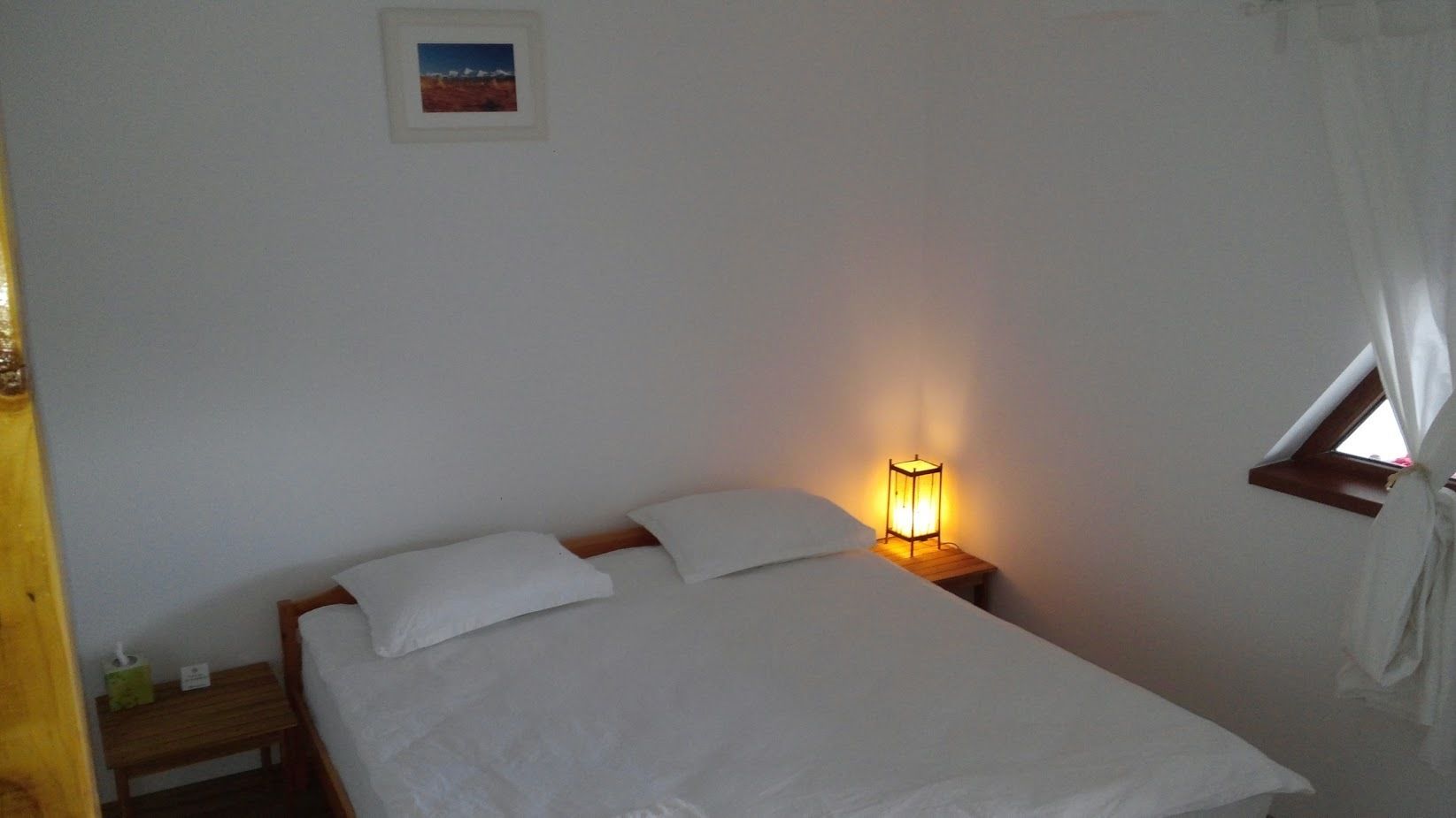 A single bed with white linens, a bedside table with a lamp, a small picture frame on the wall, and a window with curtains.