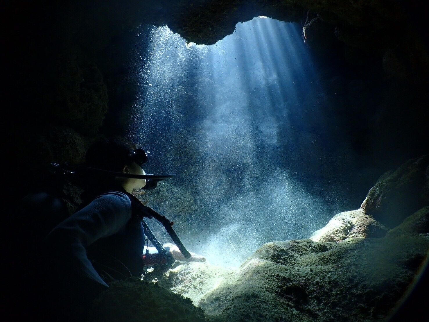 A person in scuba gear inside a cave with sunlight streaming through an opening.