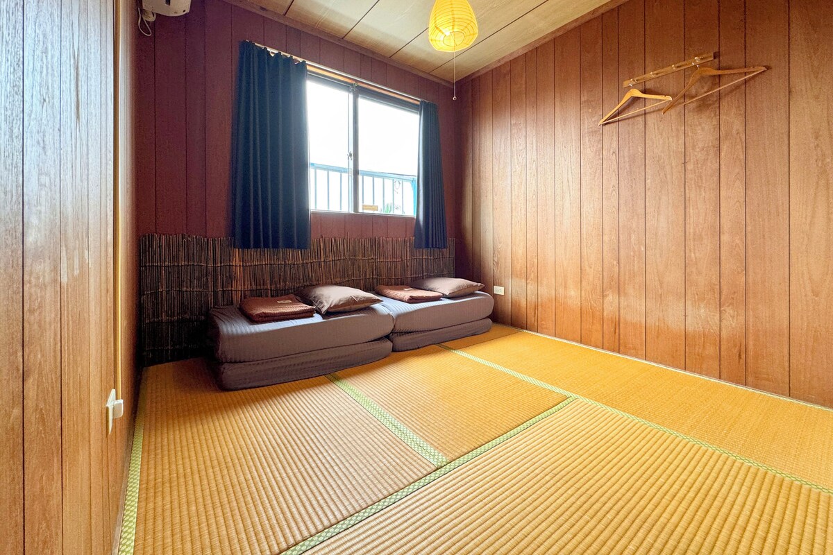 Une pièce de style japonais traditionnel, avec un sol recouvert de tatamis, un lit bas en bois agrémenté de coussins, une fenêtre avec des rideaux et un luminaire suspendu.