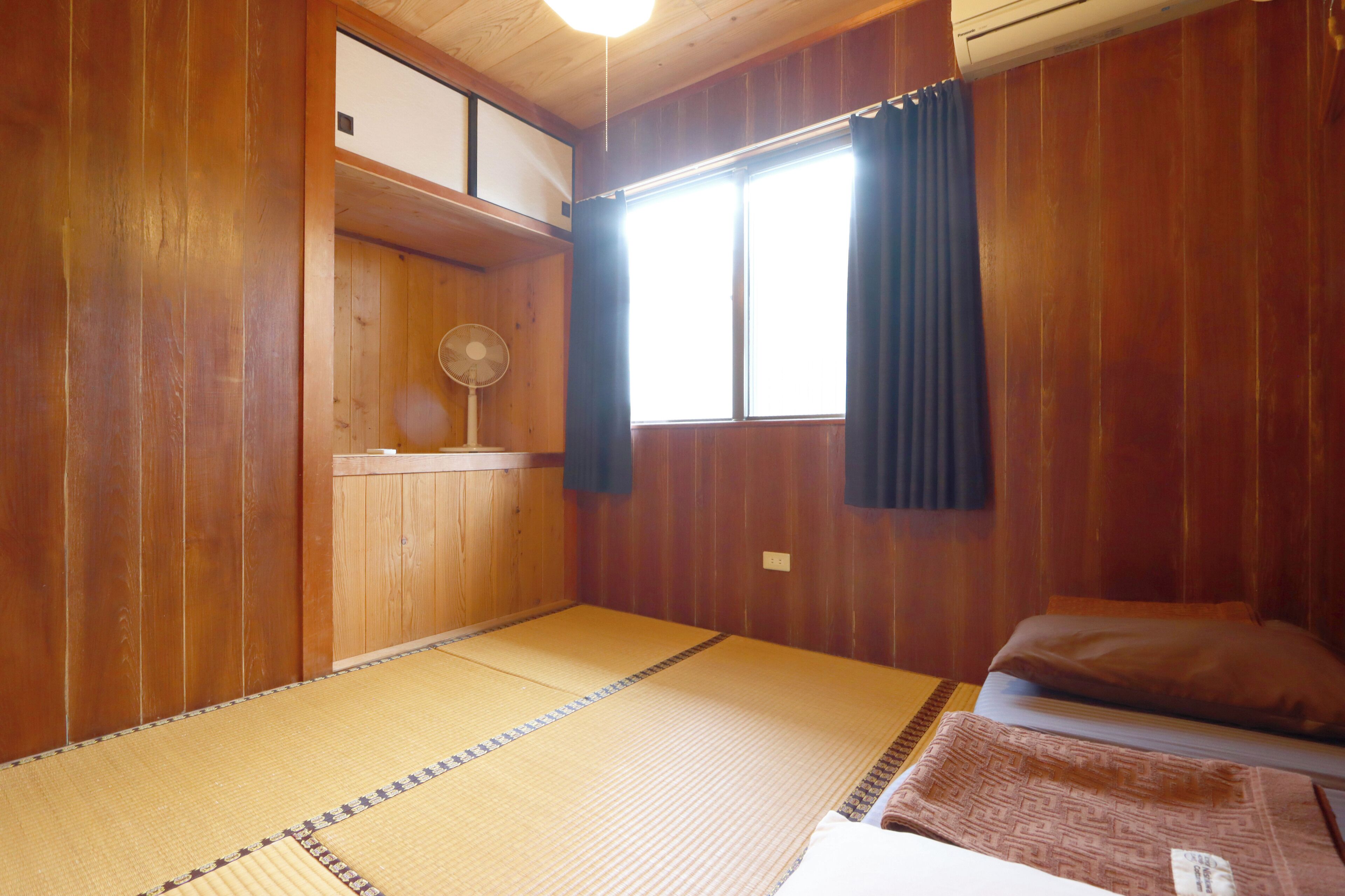 A traditional Japanese room with tatami flooring, a futon bed, a fan, and a window with curtains.