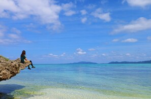 Une personne assise sur un rocher, au bord d’une mer bleue et limpide, avec des montagnes qui se dessinent à l’horizon.