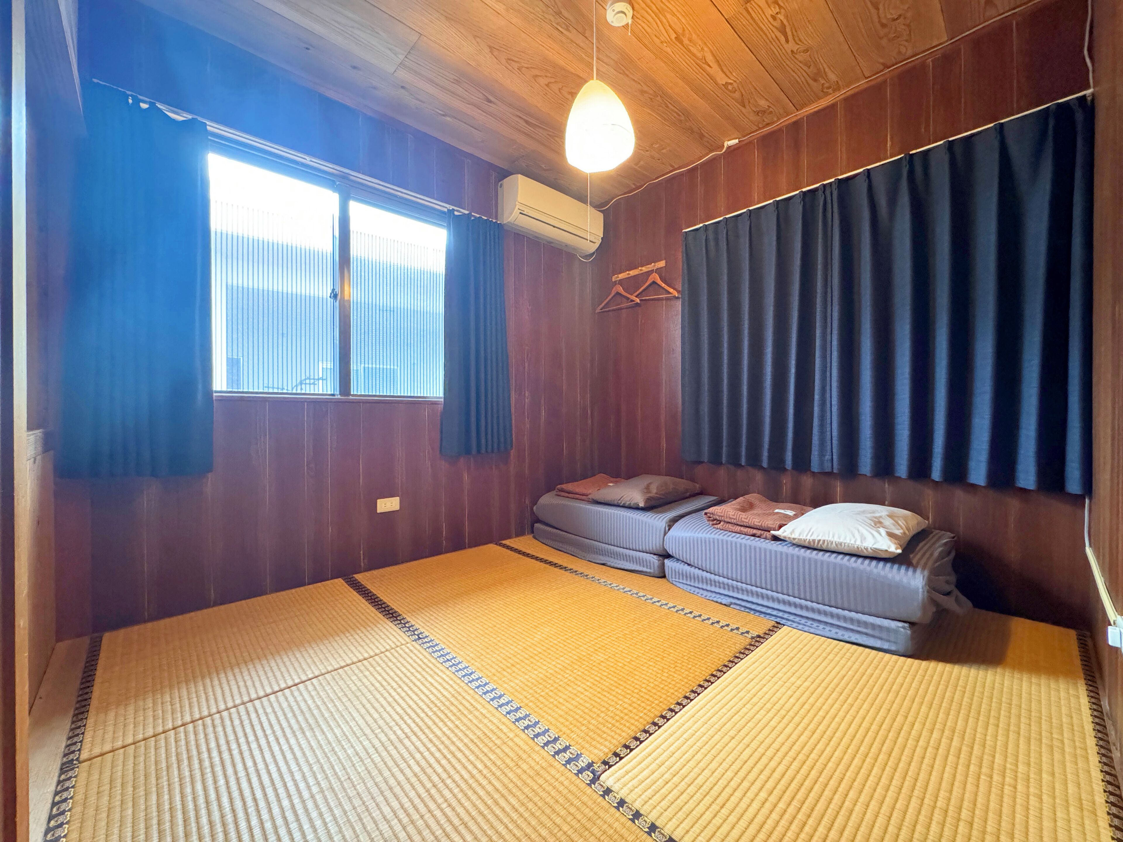 Habitació amb esteires de tatami, un llit amb almoades, una finestra amb cortines i un aparell d'aire condicionat.