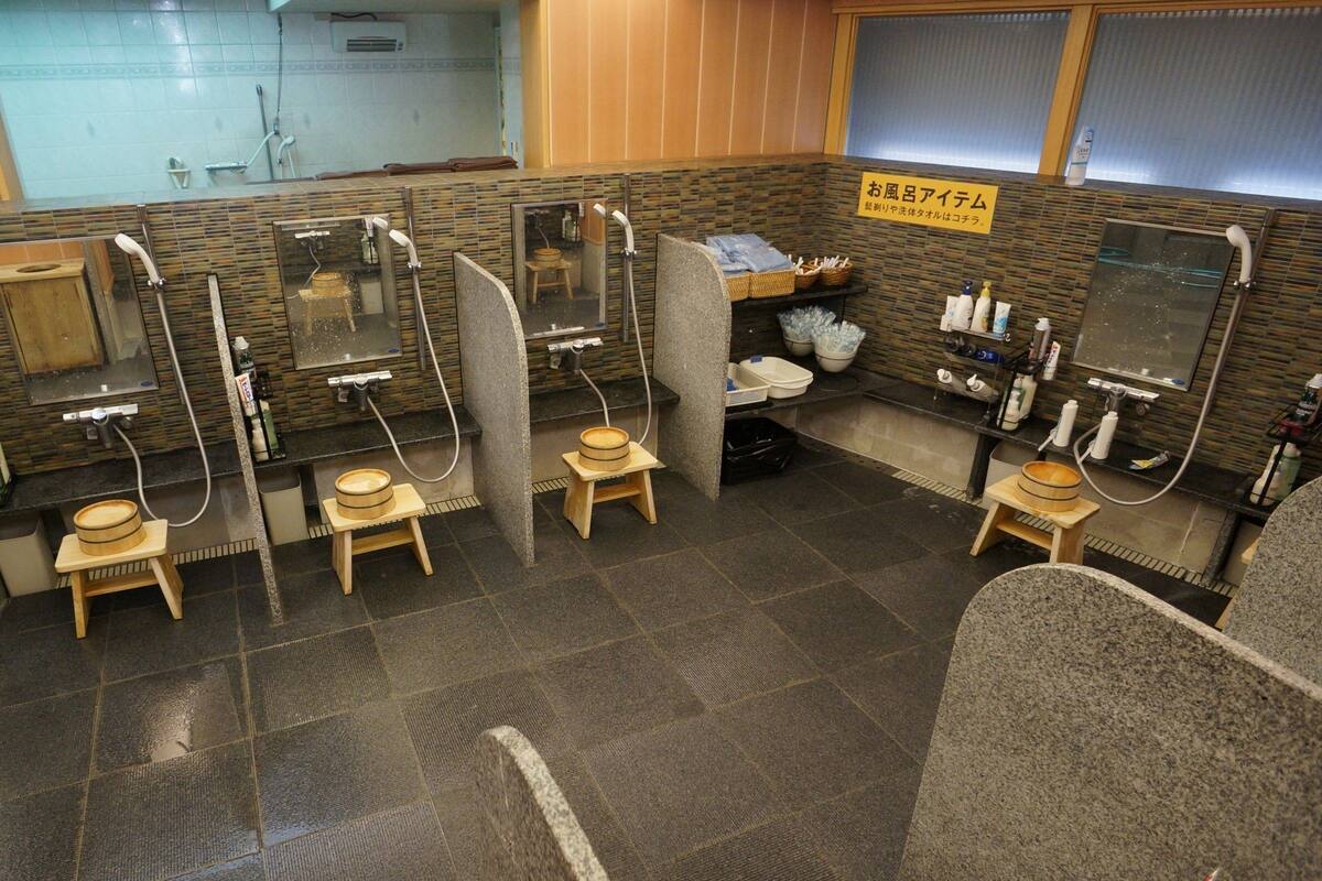 Eine öffentliche Toilette mit mehreren Kabinen, jeweils ausgestattet mit Toilette, Bidet und Handtrockner. Holzschemel und ein kleines Regal mit Toilettenartikeln sind vorhanden.