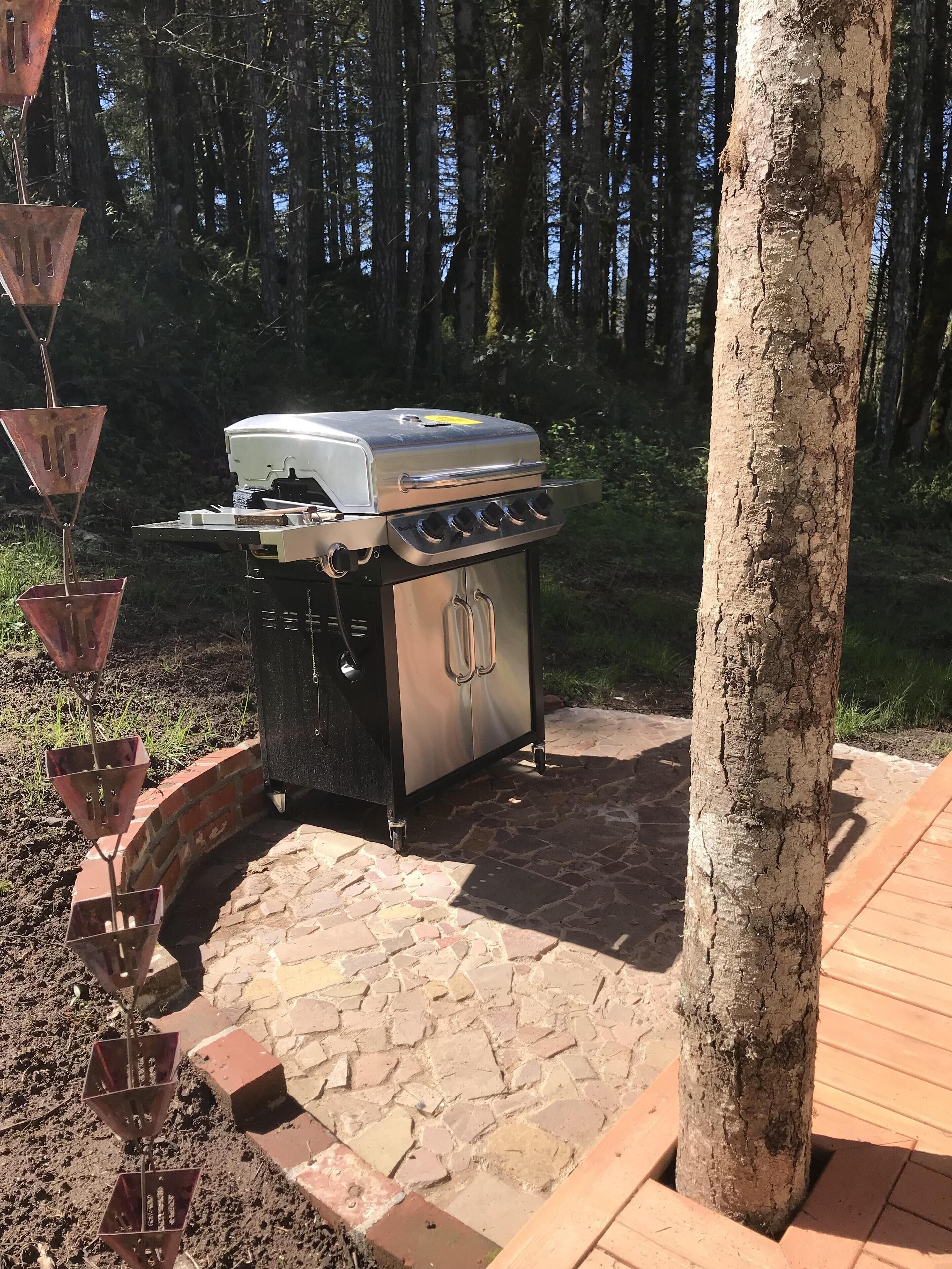 An outdoor grill with a stainless steel finish, a built-in thermometer, and a side burner. It is situated on a stone patio with a brick border, surrounded by a wooden deck and a tree.