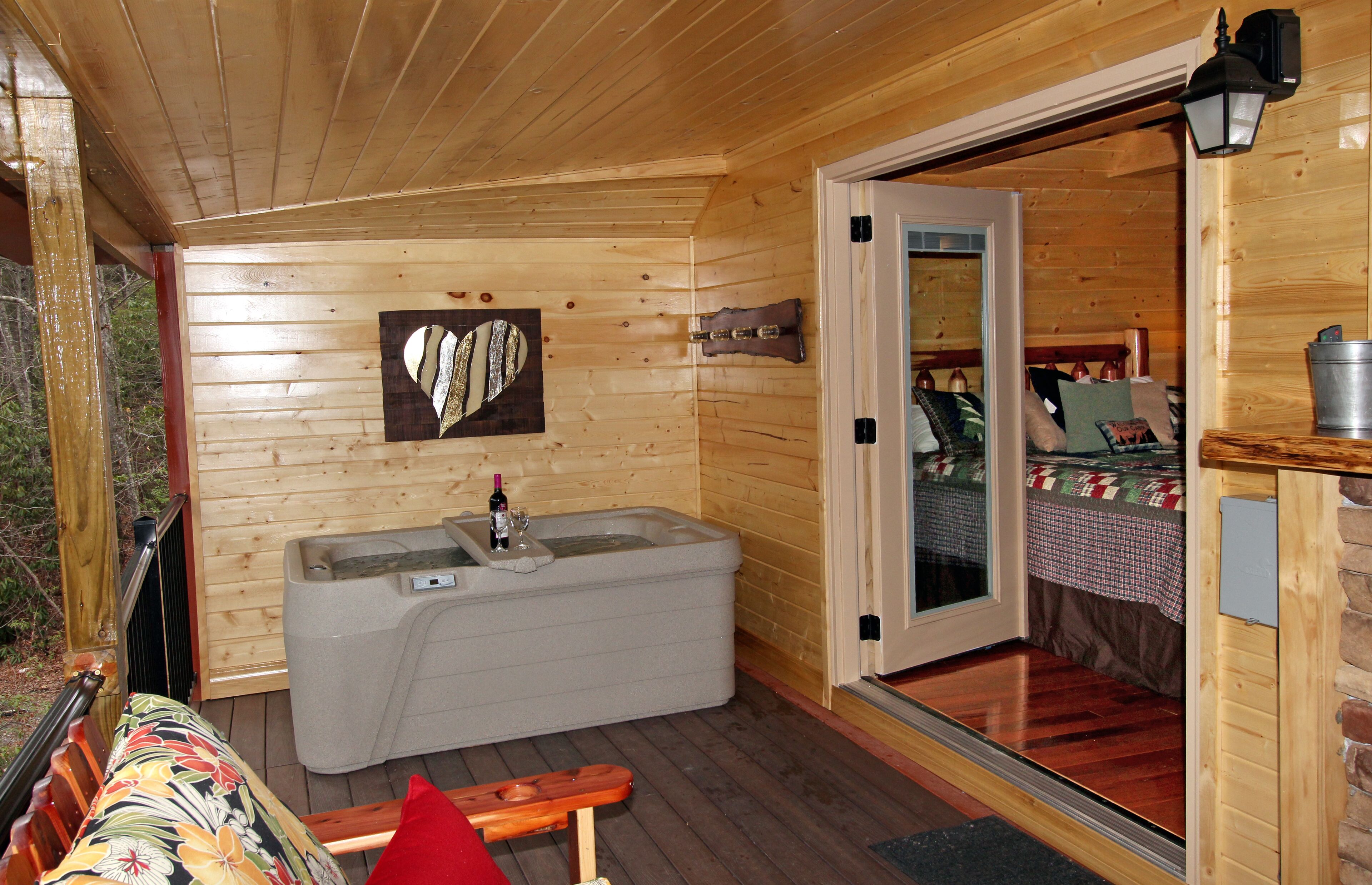 A wooden cabin with a hot tub, a bench, and a door leading to a bedroom with a bed and a quilt.