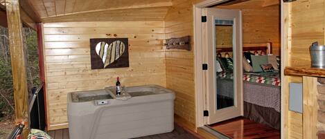 A wooden cabin with a hot tub, a bench, and a door leading to a bedroom with a bed and a quilt.