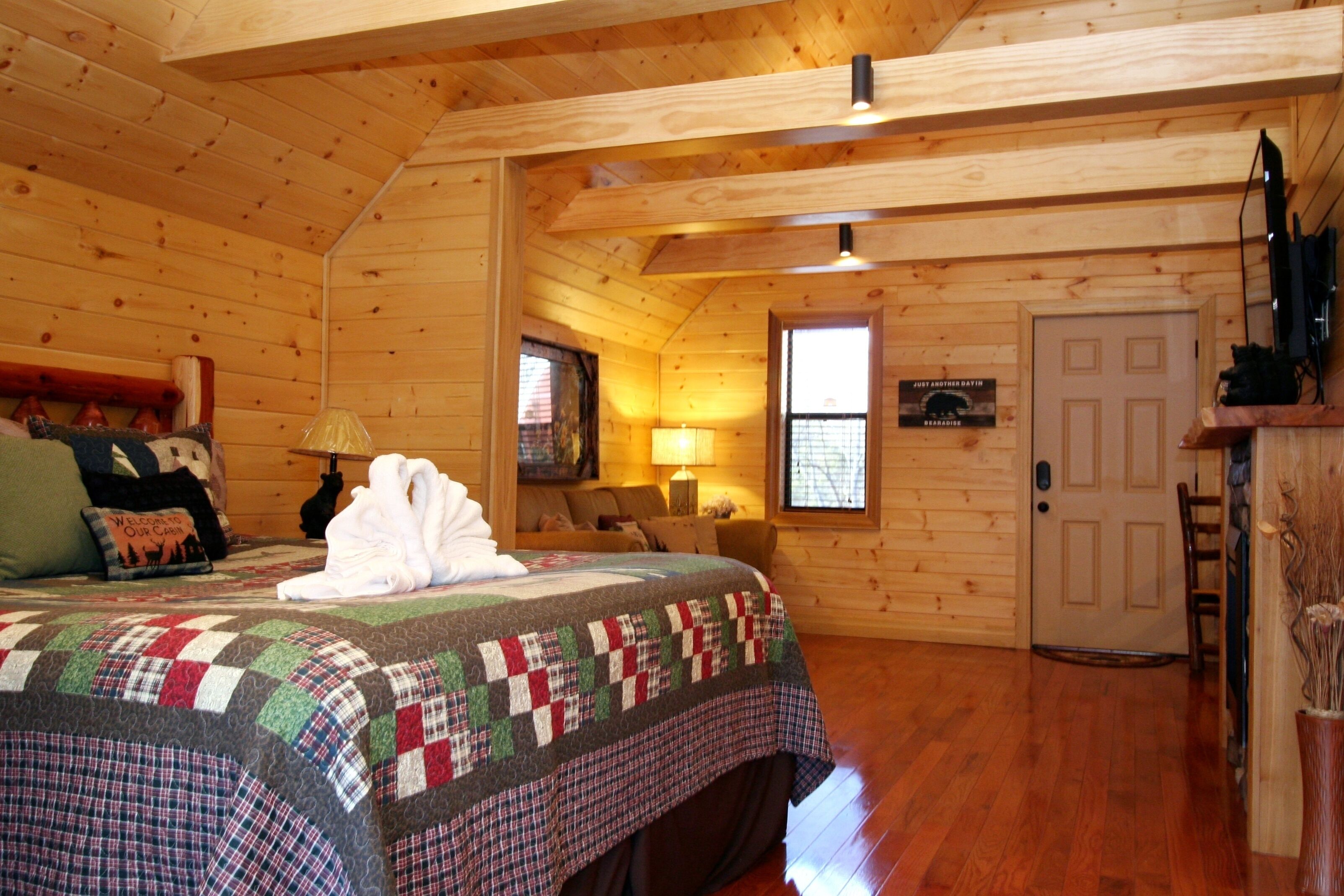 A cozy log cabin bedroom with a large bed, a wooden ceiling, a door, and a small window.