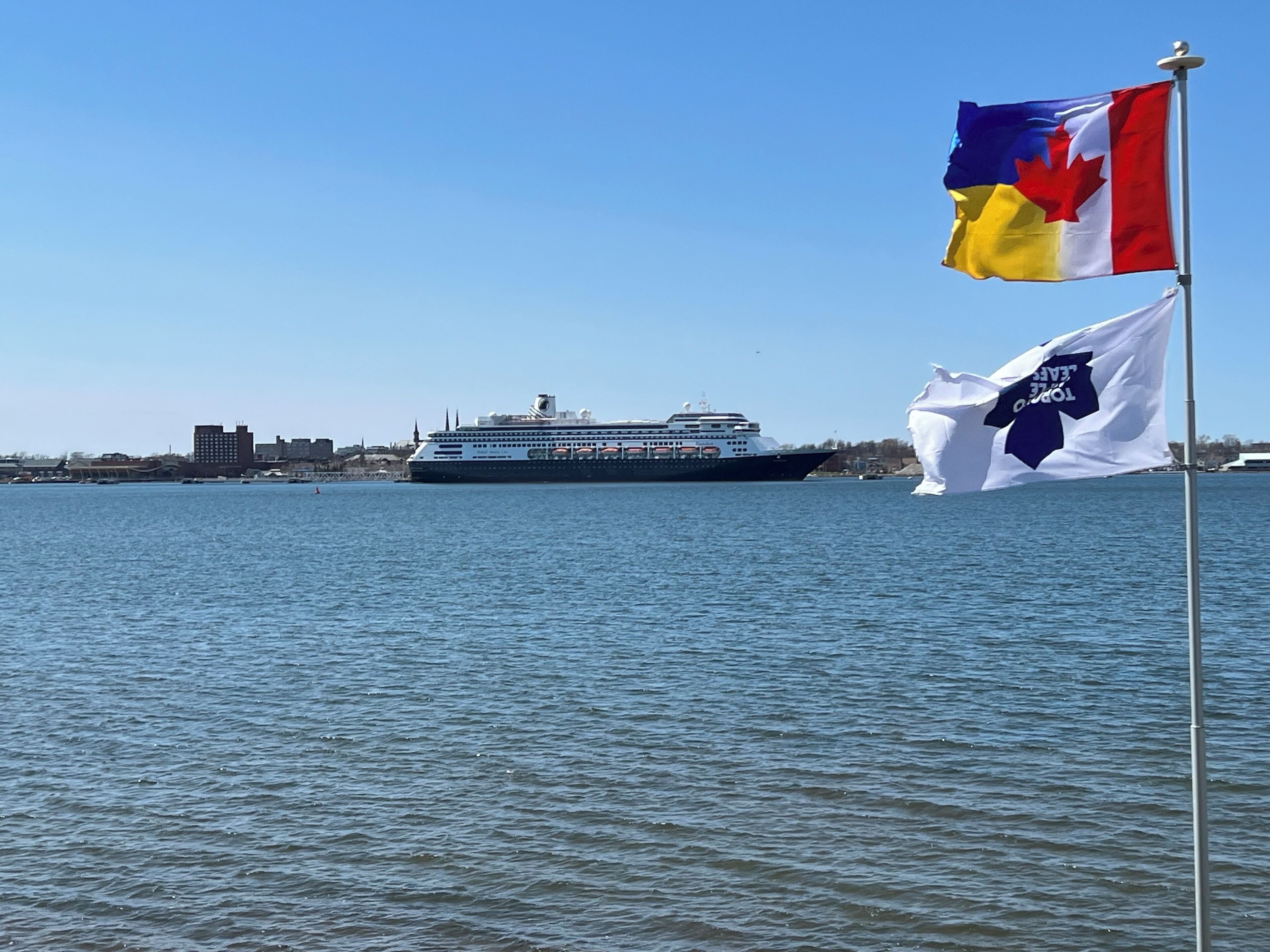 A cruise ship docked at a port with two flags, one of Canada and one of the United States, in the foreground.