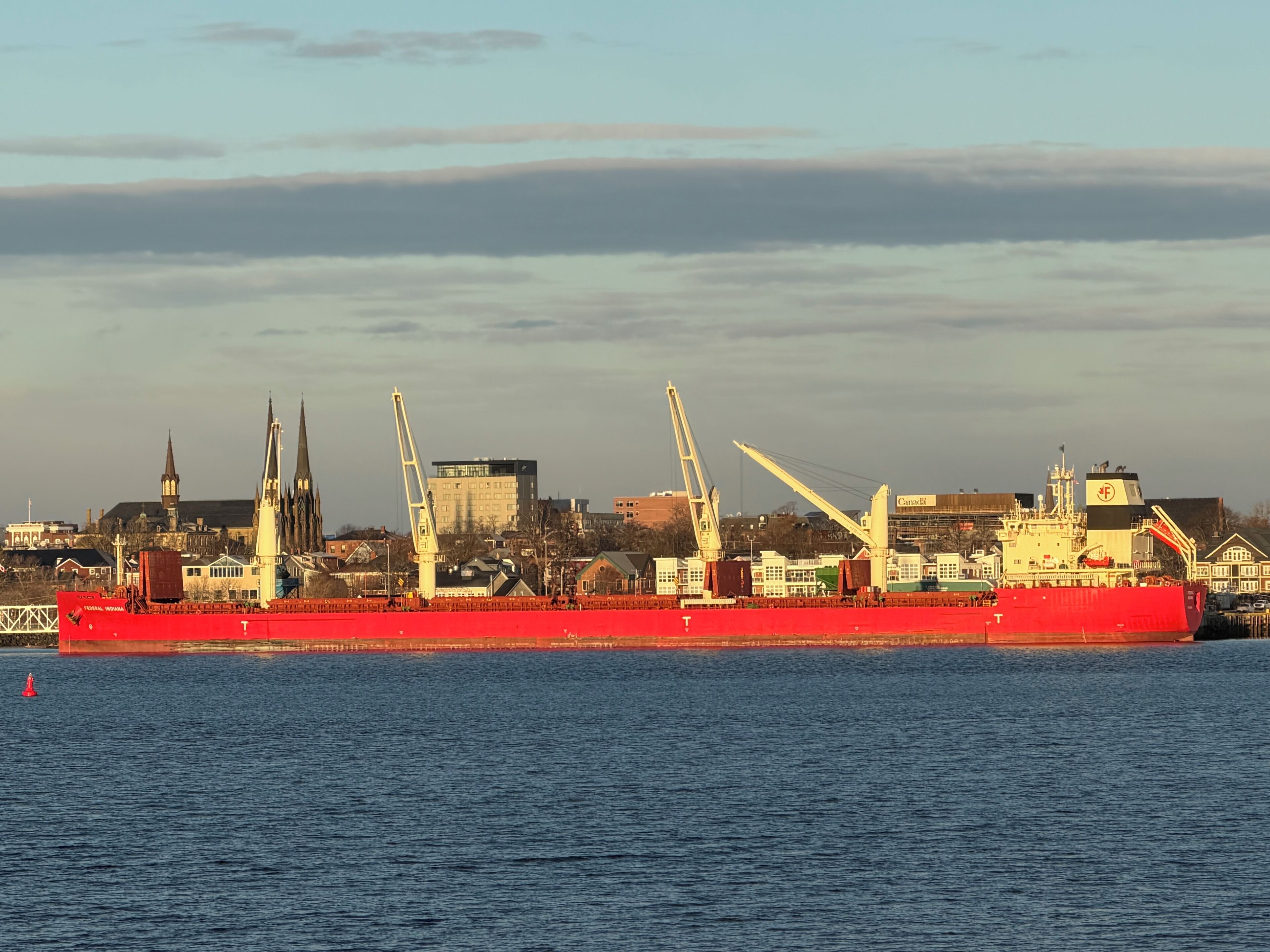 A large red cargo ship docked at a port with cranes and buildings in the background.