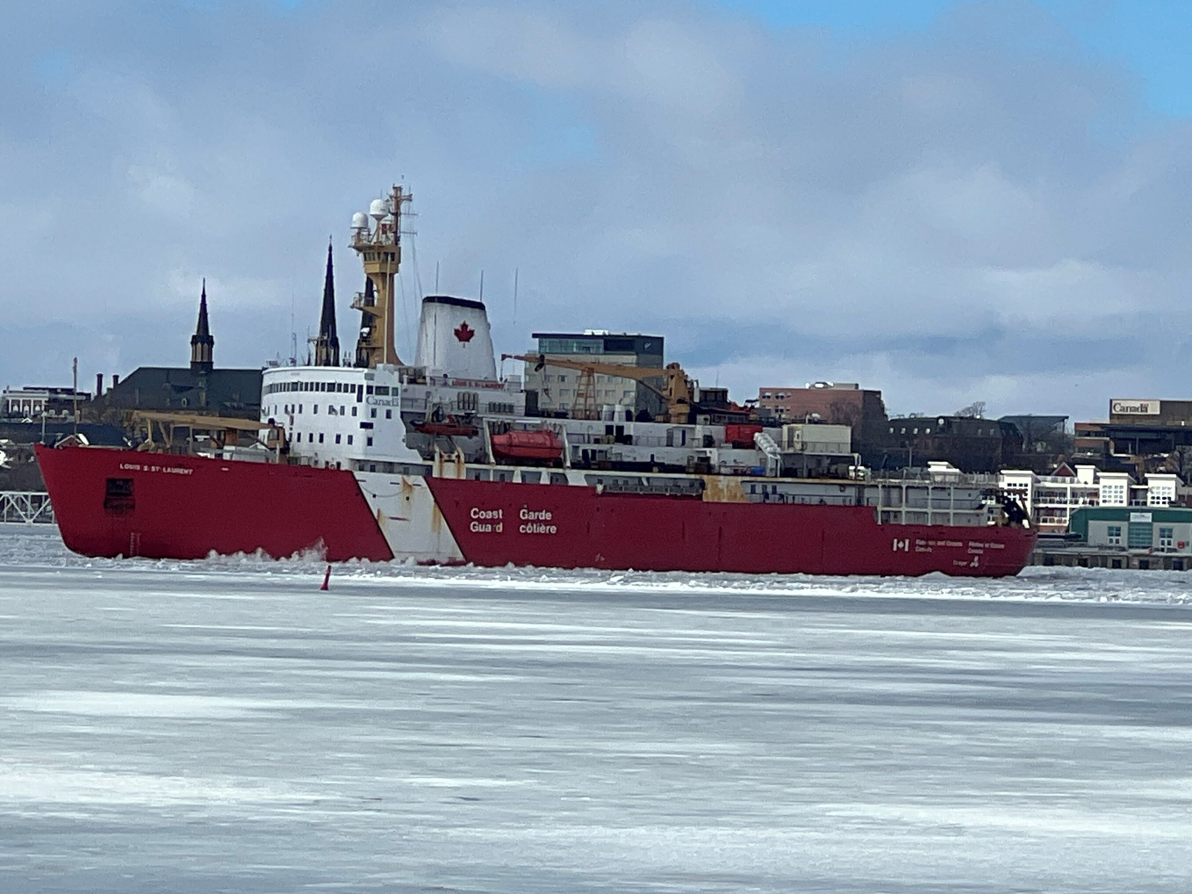 Un gran barco rojo con el nombre "Coast Guard" en su costado, navegando sobre aguas heladas con un paisaje urbano al fondo.