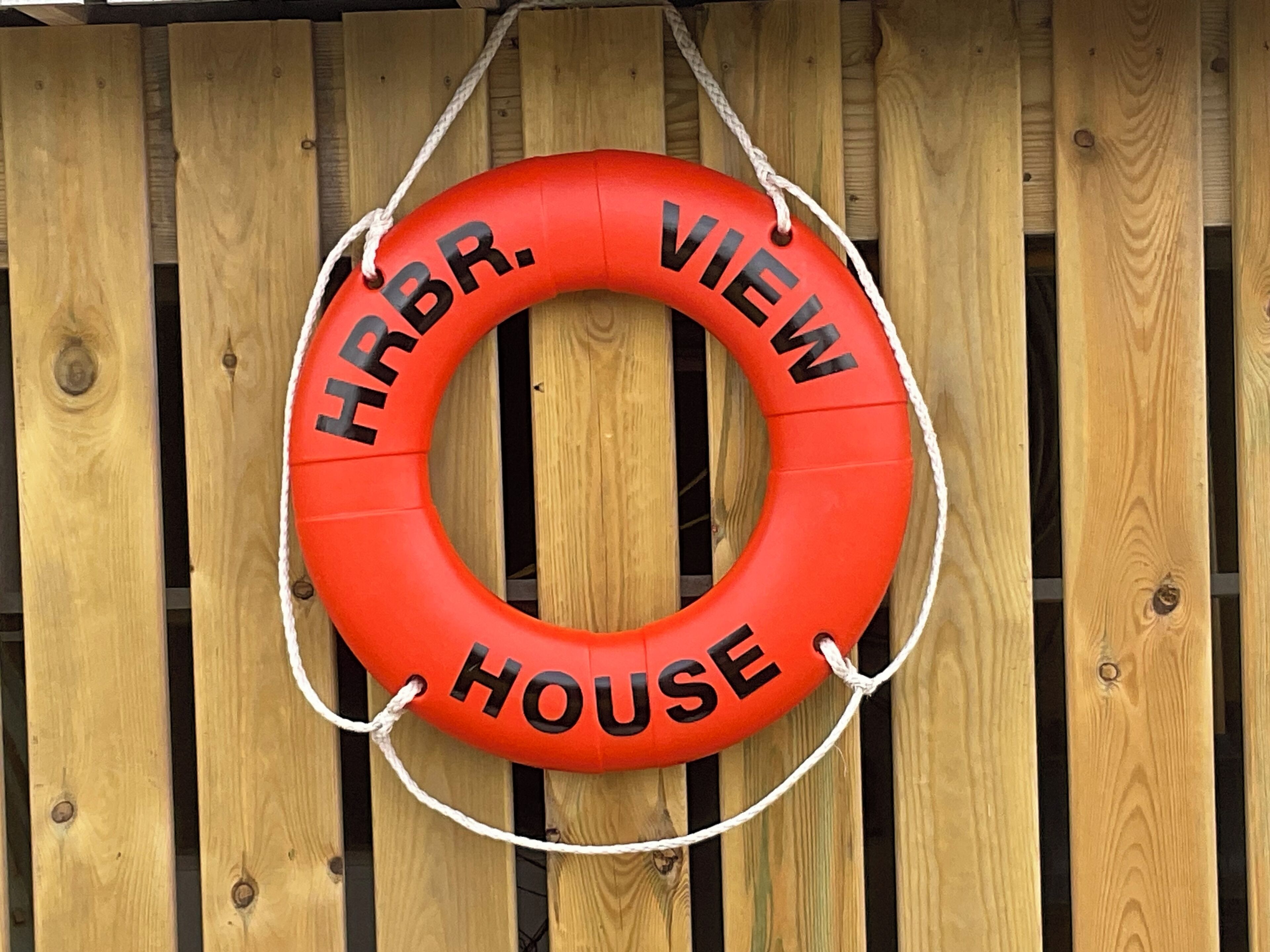 A red lifebuoy with "HRBR. VIEW HOUSE" text, hanging on a wooden fence.